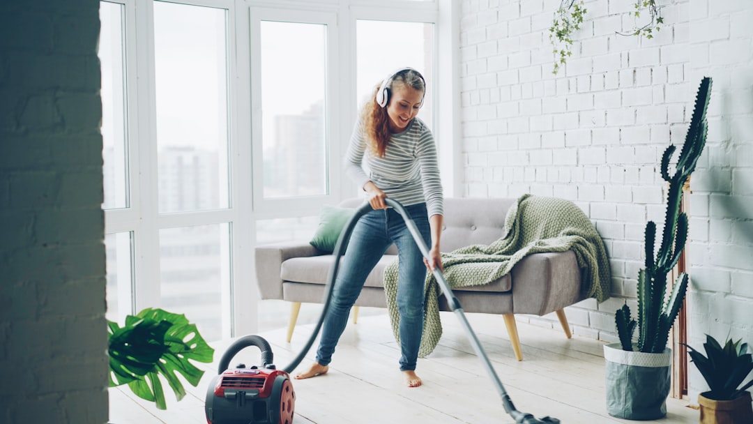 Attractive young lady is dancing and singing listening to music through wireless headphones during housecleaning at home, girl is hoovering floor and enjoying tune.